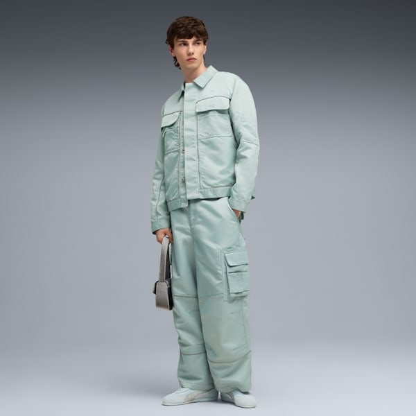 A model poses against a grey backdrop, wearing a light green outfit and holding a bag.