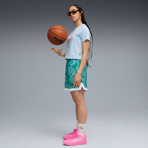 Against grey, a model wearing sunglasses, a t-shirt, patterned shorts and pink trainers is holding a basketball.