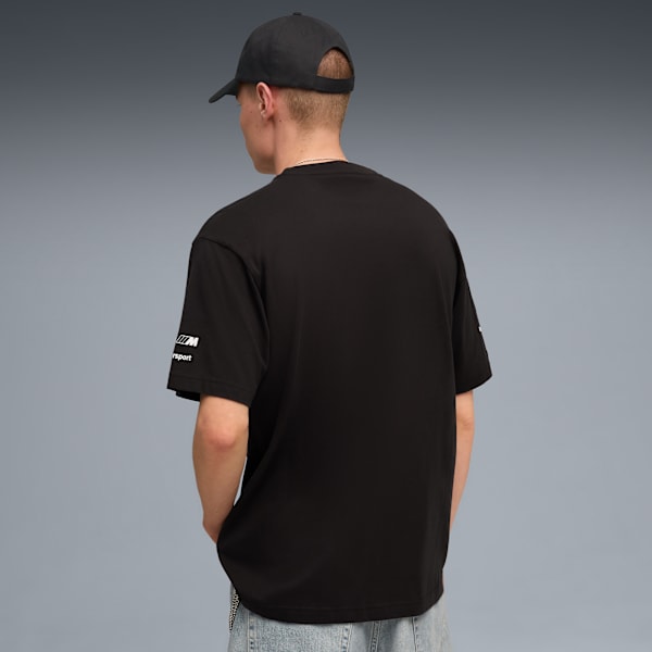 A model faces away wearing a black tee and cap against a grey backdrop.