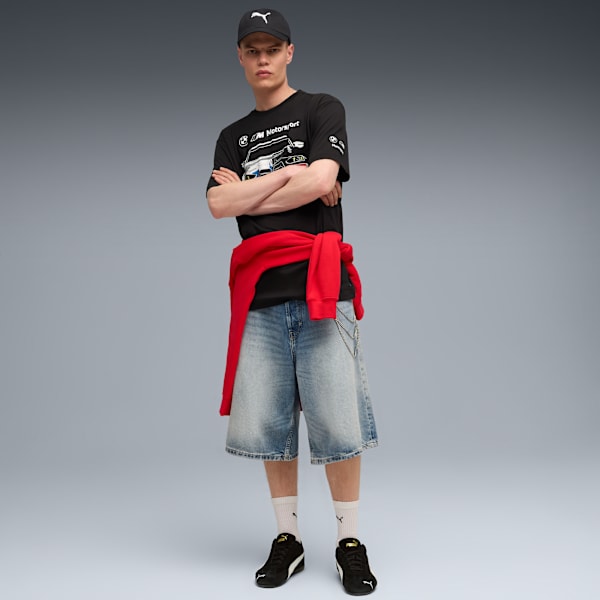 On grey, a model stands with crossed arms, wearing a black tee, denim shorts and black trainers.