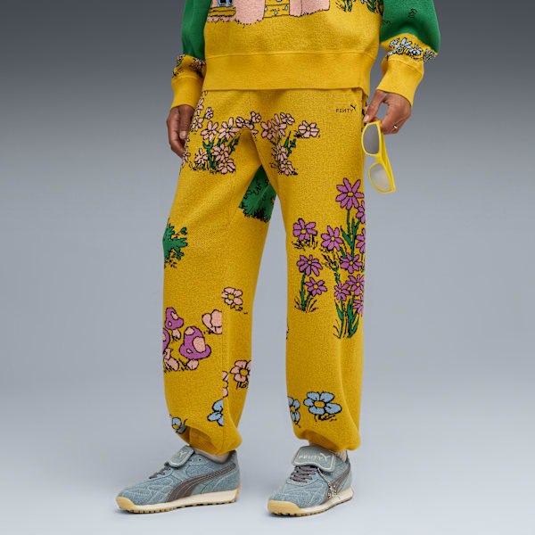 On a grey backdrop, a model wears floral yellow trousers and trainers, holding sunglasses.