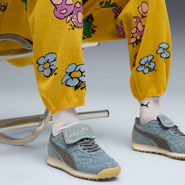 On a light backdrop, someone sits wearing floral trousers and blue Fenty x Puma trainers.