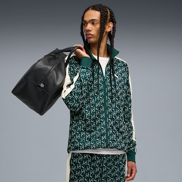 A model with braids holds a black bag, wearing a patterned green tracksuit against grey.