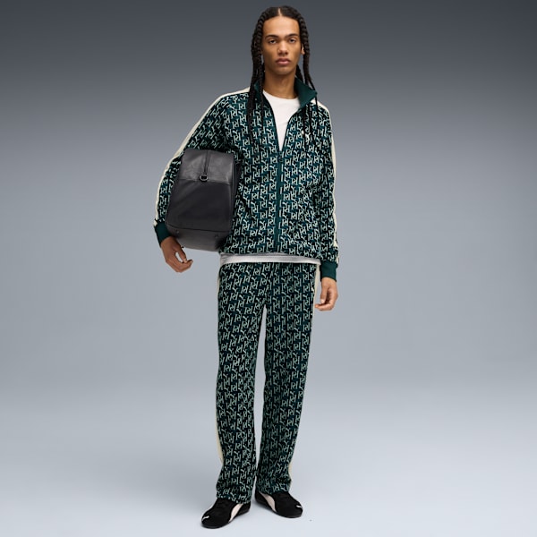 Against grey, a model holds a black bag, wearing a patterned green tracksuit and black trainers.