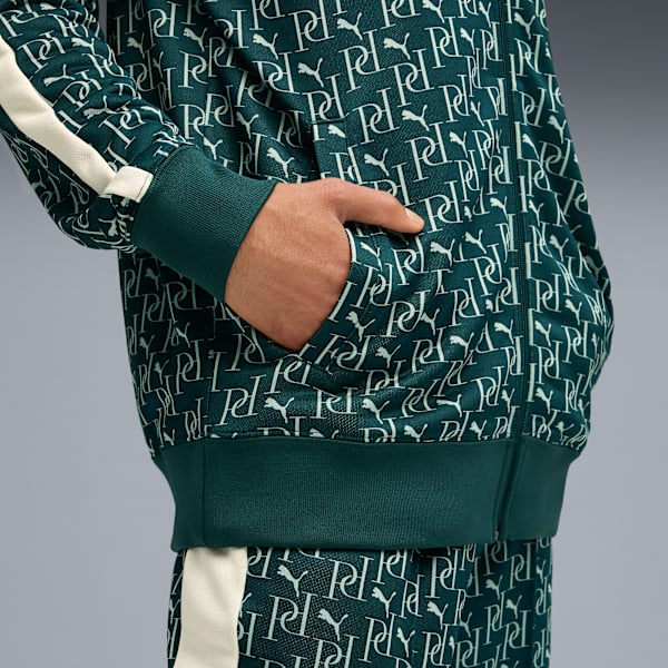 A person with hand in pocket wearing a green tracksuit with a white pattern against a grey background.