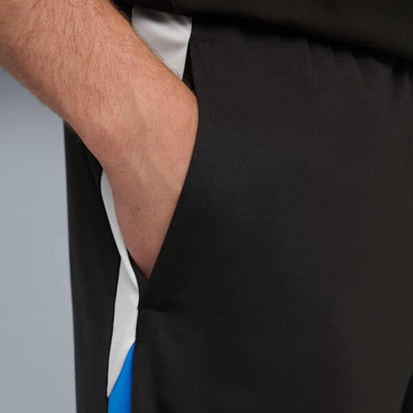 A hand in the pocket of black shorts with white and blue stripes on a grey backdrop.