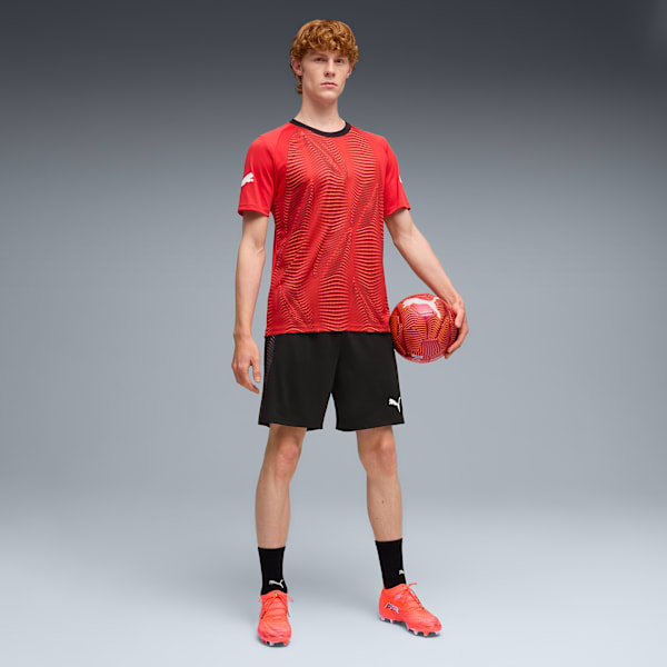 Against grey, a model poses with a ball, wearing a red t-shirt, black shorts and orange shoes.