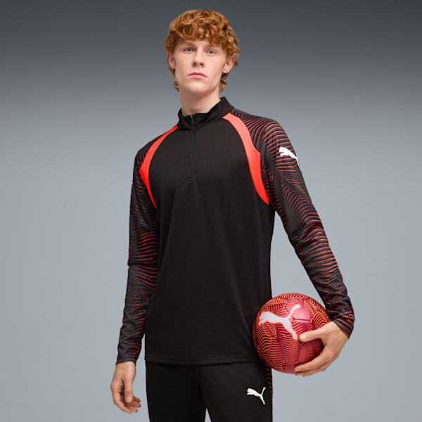 Against grey, a model holds a patterned ball, wearing a black and red long-sleeved top.