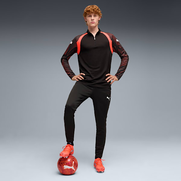 On grey, a model poses with a foot on a soccer ball, wearing a black and red tracksuit.