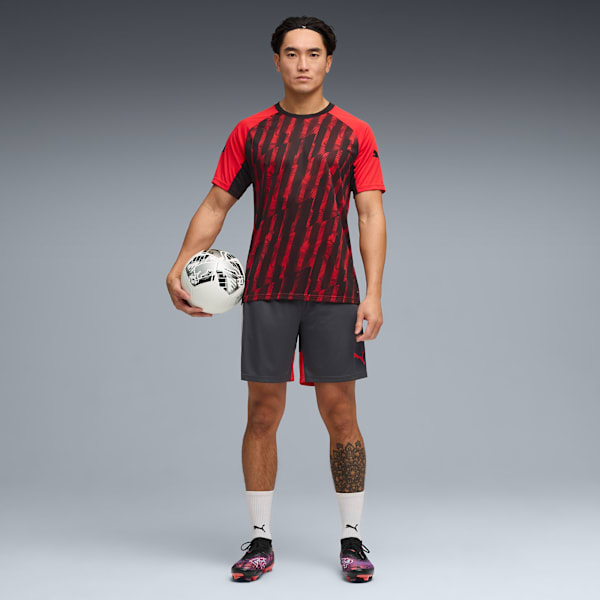 Against grey, a model holds a football, wearing a red and black shirt, grey shorts and football boots.