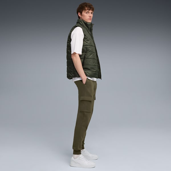 Against grey, a model poses in a green gilet, white t-shirt, green trousers and white trainers.