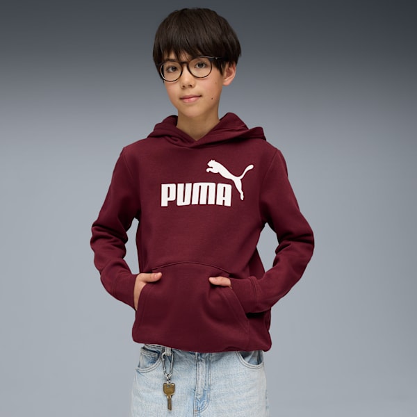 A child with glasses poses in front of a grey background wearing a burgundy Puma hoodie.