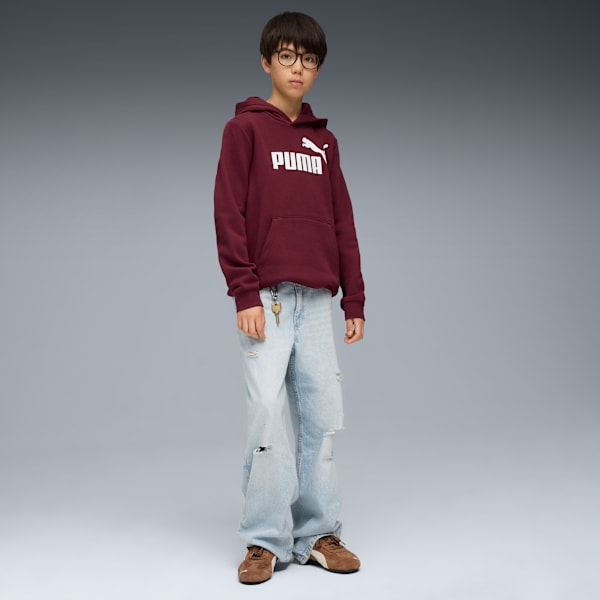 A boy with glasses stands against a grey backdrop, wearing a maroon hoodie, ripped jeans and trainers.