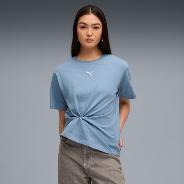A model poses against grey wearing a blue t-shirt with a knot and brown trousers.
