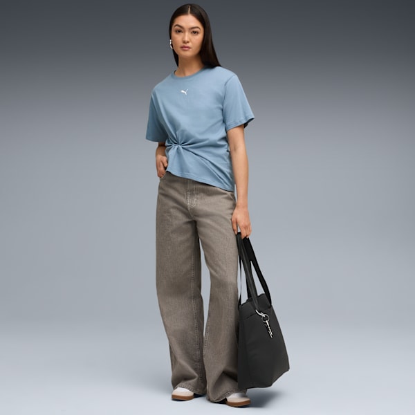 A model stands against grey, wearing a blue t-shirt, wide jeans and holding a black bag.