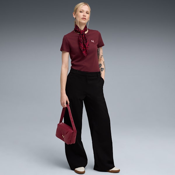 A model poses against a grey backdrop, wearing a maroon t-shirt, black trousers and holding a red bag.