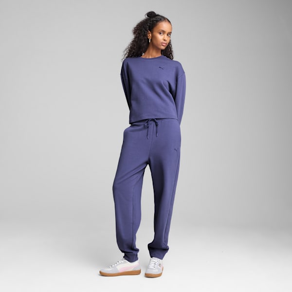 A model stands against a grey backdrop, wearing a blue sweatshirt and matching jogging bottoms.