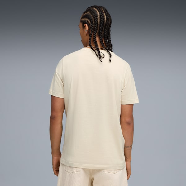 A model with braided hair faces away, wearing a cream t-shirt and shorts against a grey backdrop.