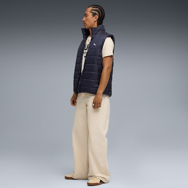 Against grey, a model stands wearing a navy vest, cream tee, wide trousers, and trainers.