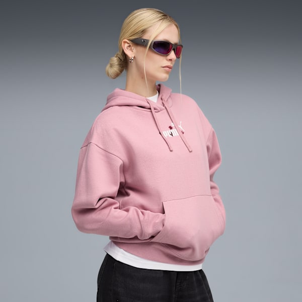 Against a grey backdrop, a model poses in a pink hoodie, jeans, and sunglasses.