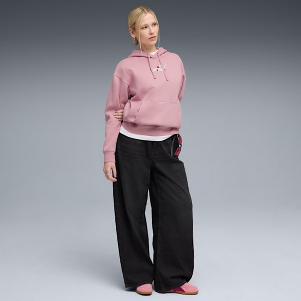 A model stands against grey wearing a pink hoodie, black jeans, and pink shoes.