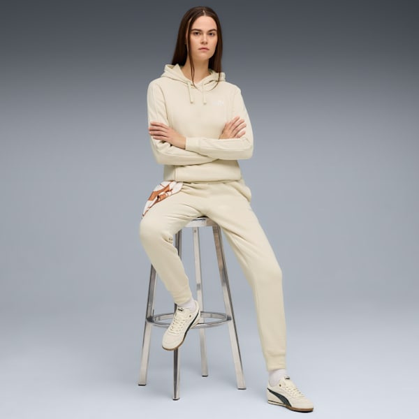 On grey, a model sits with arms crossed, wearing a cream hoodie and jogging bottoms.