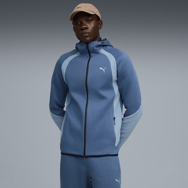 A model stands against grey, wearing a blue hooded jacket, trousers, and a beige cap.