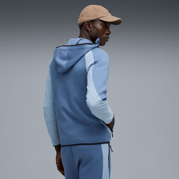 Against grey, a model glances left, wearing a cap and blue hooded tracksuit with hands in pockets.