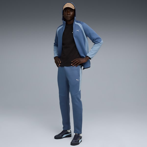 Against a grey backdrop, a model poses in a blue tracksuit and dark trainers.