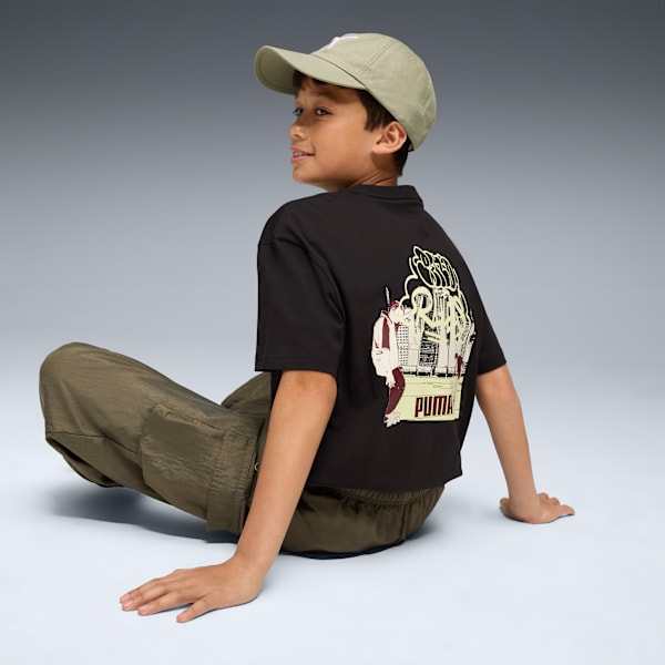 A child sits against a grey backdrop, wearing a cap, printed tee, and trousers.