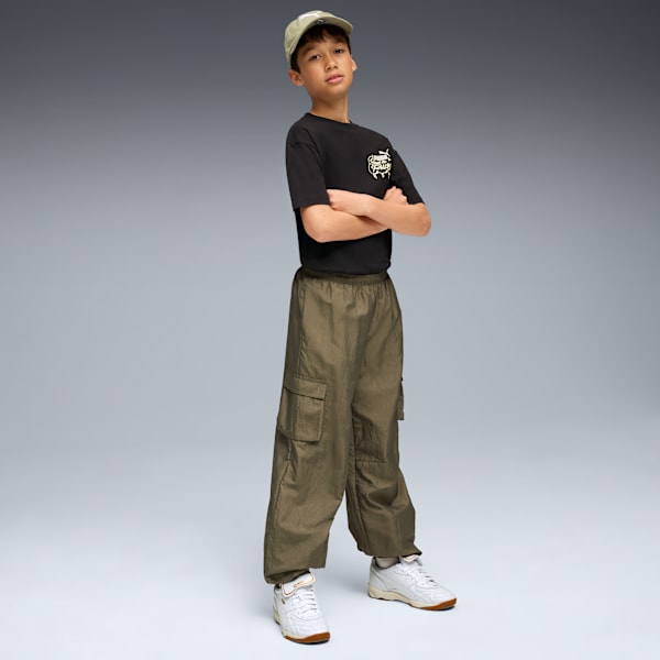 A model with arms crossed stands in front of a grey background wearing a black t-shirt and green trousers.