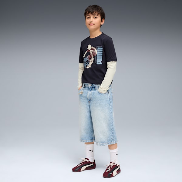 A model stands on a grey backdrop wearing a graphic tee, denim shorts and trainers.