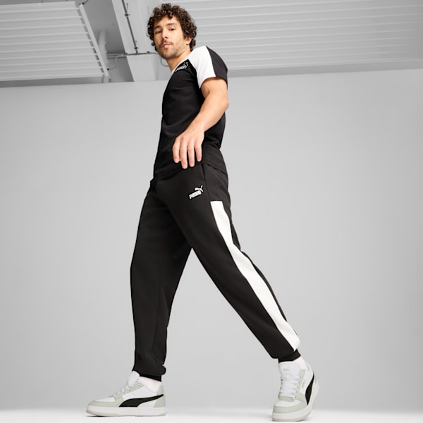 A model is walking against a grey backdrop, wearing a black and white t-shirt, trousers and trainers.