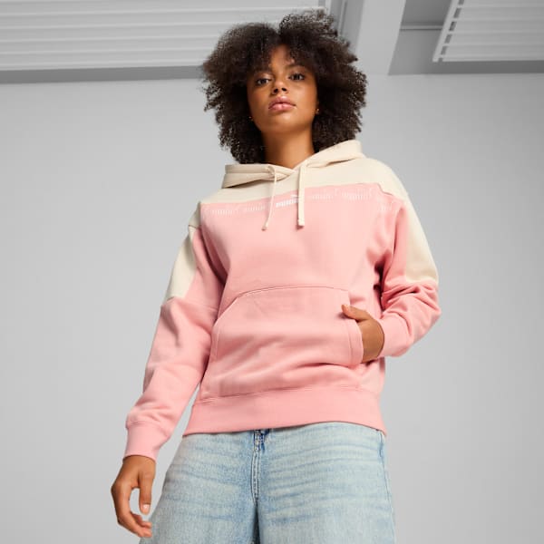 A model poses against a grey backdrop, wearing a pink and cream hoodie and denim jeans.