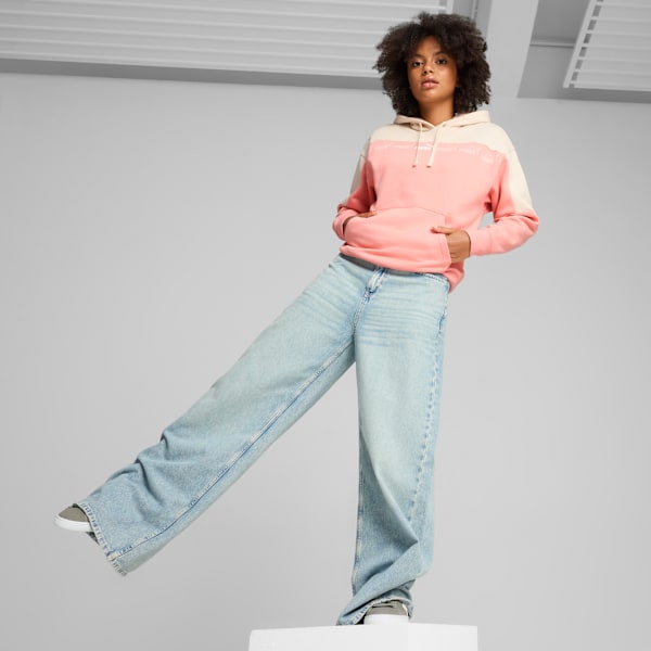 A model poses on grey in a pink hoodie and wide jeans with one leg extended.