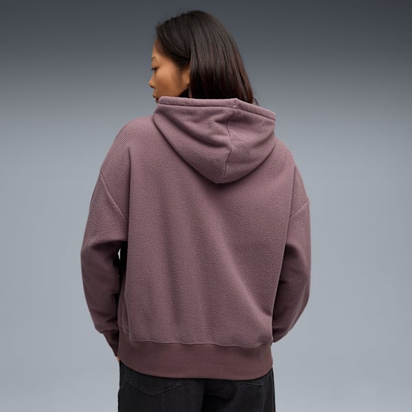A model faces away, wearing a textured mauve hoodie and dark trousers against grey.