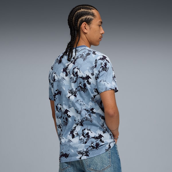 Against grey, a model with braids wears a patterned shirt and jeans, looking away.