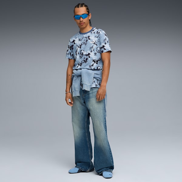 A model in front of a grey backdrop wearing a patterned t-shirt, jeans and blue glasses.
