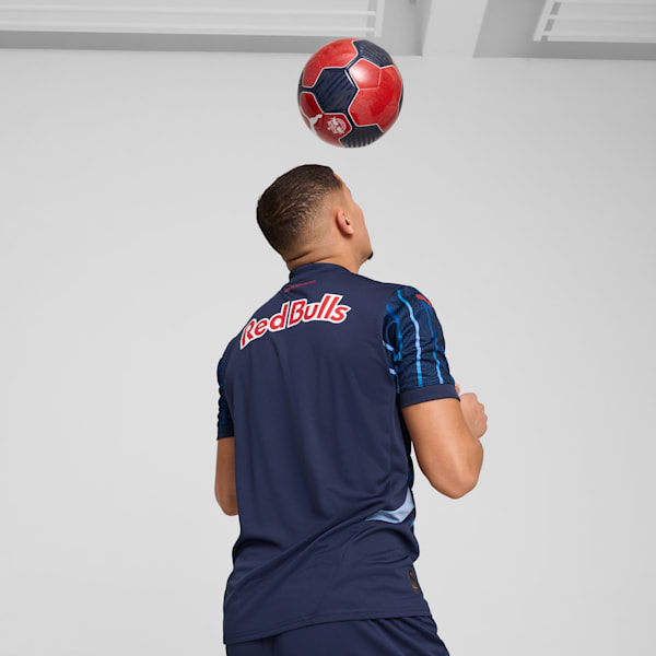 Against a grey backdrop, a model in a blue shirt heads a red and navy football.