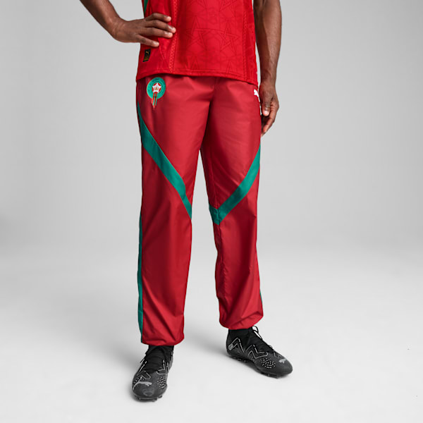 On grey, a model wears red trousers with green stripes and black running shoes, posing.