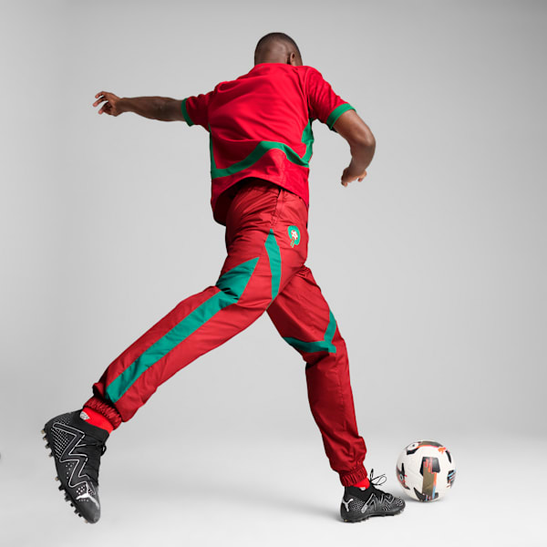 A model kicks a football in a red and green kit, with running shoes, against a grey backdrop.