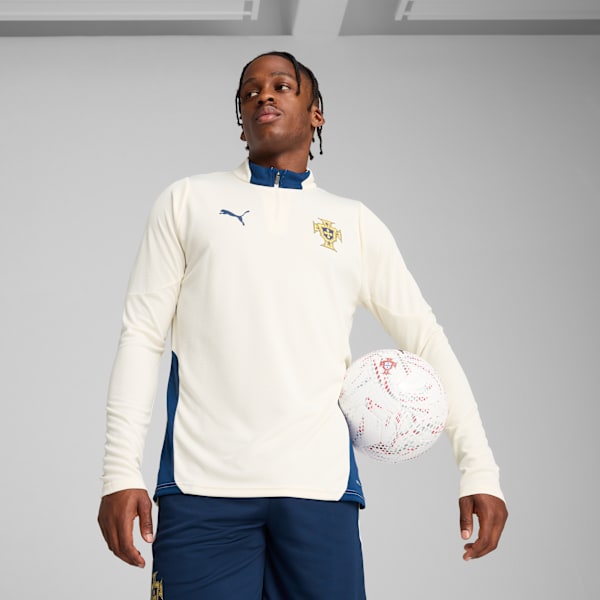 On grey, a model holds a football, wearing a cream top and blue shorts.