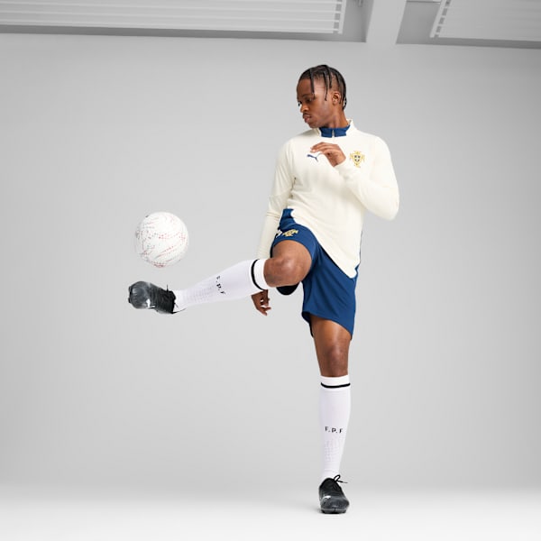 On grey, a model kicks a football wearing a cream top, navy shorts, and black football boots.
