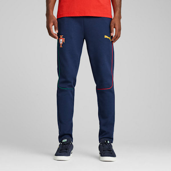 Against grey, a model wears navy joggers, a red top, and trainers, standing still.