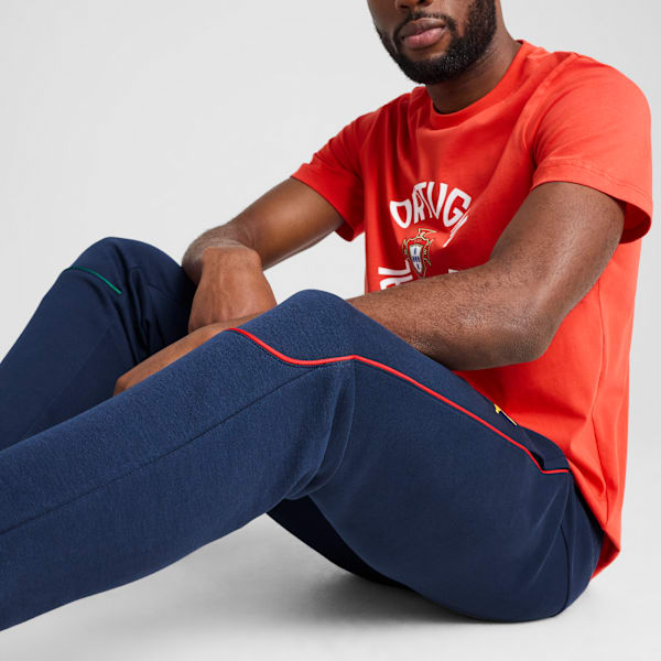 On a grey backdrop, a model poses seated, wearing a red t-shirt and navy tracksuit bottoms.