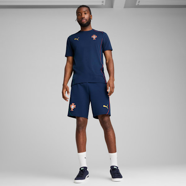 Against grey, a model stands wearing a navy t-shirt, shorts and trainers with white socks.