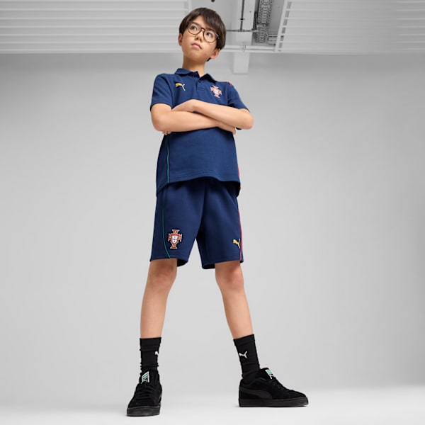 Against grey, a child models a dark blue polo and shorts with black trainers and socks.