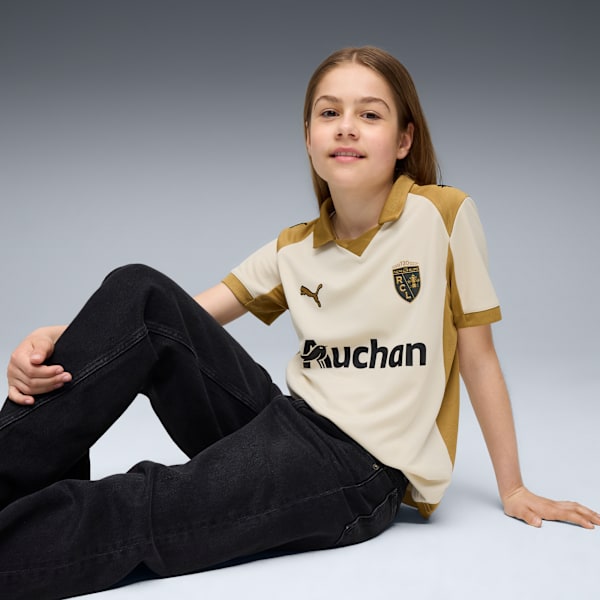 A model sits against a grey backdrop, wearing a cream and gold shirt, and dark jeans.