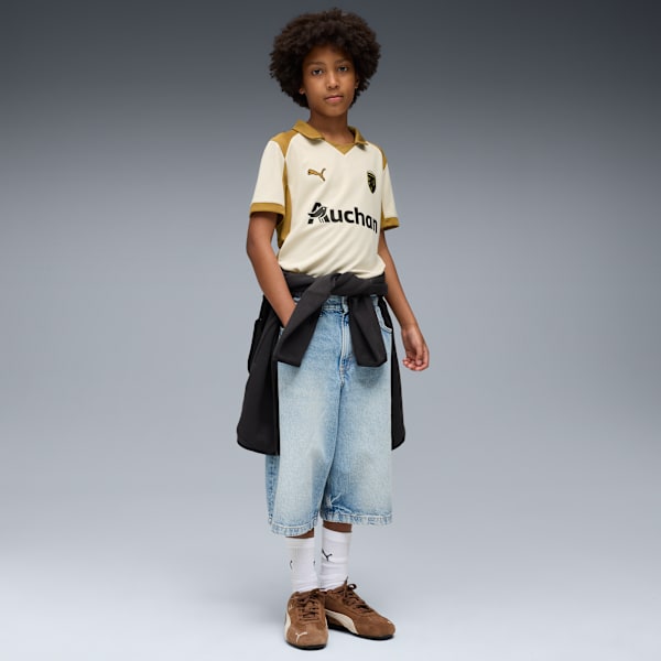 A model stands on a grey backdrop wearing a cream shirt, denim shorts and brown trainers.