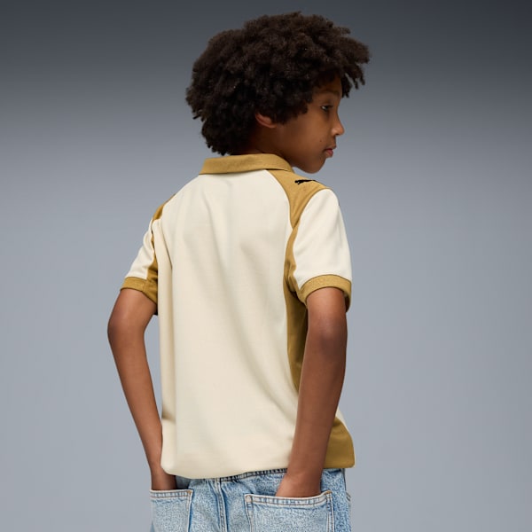 A child with hands in jean pockets poses in a cream and tan collared shirt against grey.